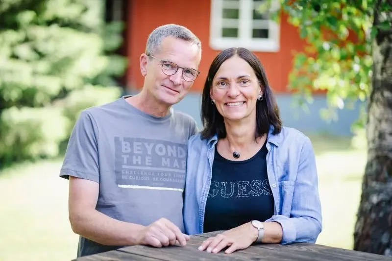 Rainer und Nicole nebeneinander an einem Tisch vor dem roten Haus